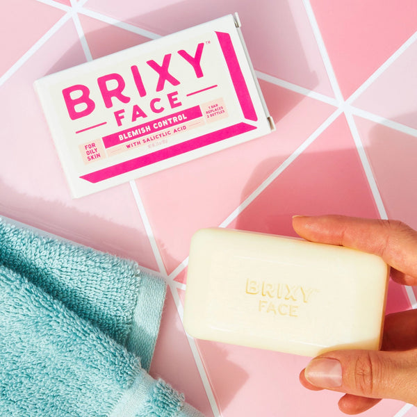 Brixy Face soap bar with packaging and a hand holding it on a pink geometric background