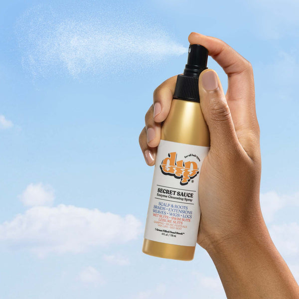 Hand holding a gold and white spray bottle against a blue sky background