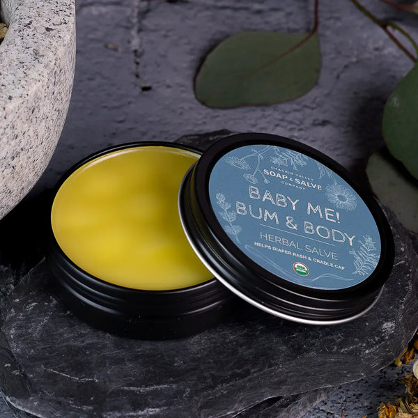 Jar of 'Baby Me! Bum & Body' herbal salve with a blue label on a stone surface.