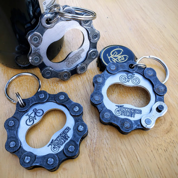 Three bicycle chain keychain bottle openers on a wooden surface with 'Tinkering Cyclist' branding.