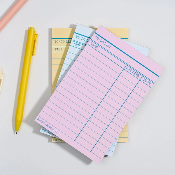 A stack of pastel notepad sheets with lined to-do lists, styled with a yellow pen, a white pen, and a peach-colored pencil on a light gray background.