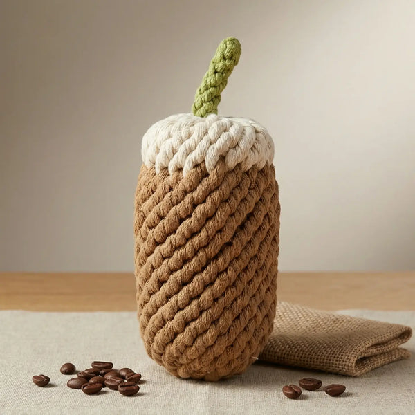 Knitted rope coffee cup toy made to look like an iced coffee. White on top with a brown body  a green 'straw' on a wooden table with coffee beans next to it for display