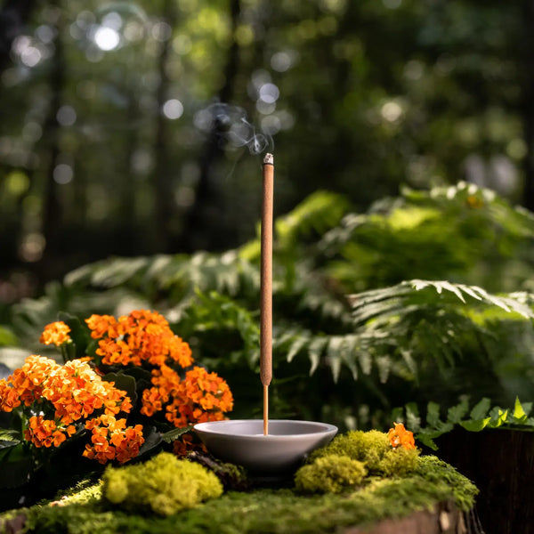 Incense stick burning in a small dish with greenery and flowers in the background