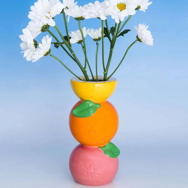 Colorful vase with stacked orange, pink, and yellow sections holding white flowers against a blue gradient background.