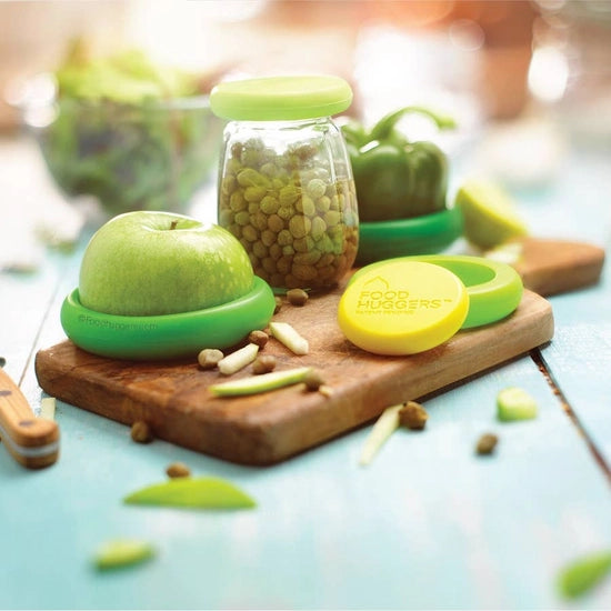 food huggers each covering half of a granny smith apple, a green bell pepper, and a jar of lentils, all displayed on a cutting board