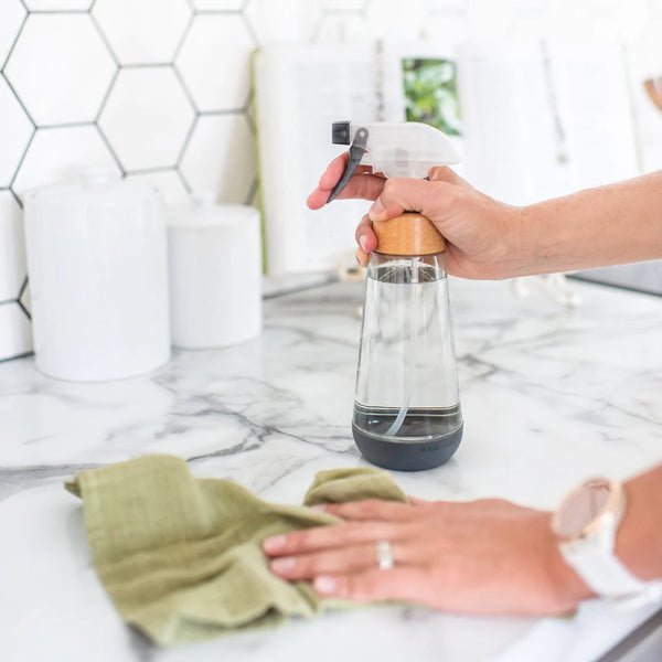 someone holding a clear glass spray bottle with silicone bottom, with a washcloth in the other hand wiping down a kitchen countertop.