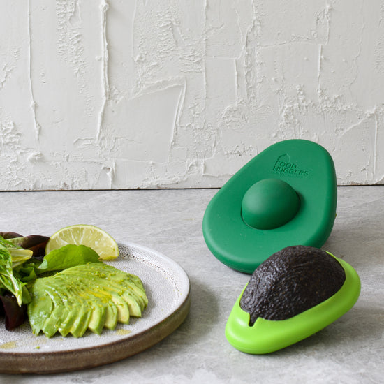 avocado huggers on a countertop next to a white hall with a plate of sliced avocado, salad greens and citrus next to them